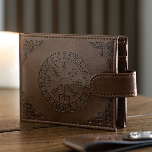 A brown leather Viking Compass wallet with a snap fastening, embossed with Norse symbols and a circular Vegvisir motif, rests on a wooden surface with a blurred candle in the background.