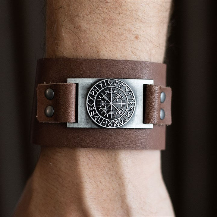 A close-up of a person’s wrist adorned with a wide brown leather cuff Viking bracelet, featuring metal rivets and a circular metal plate engraved with runic symbols and intricate designs.