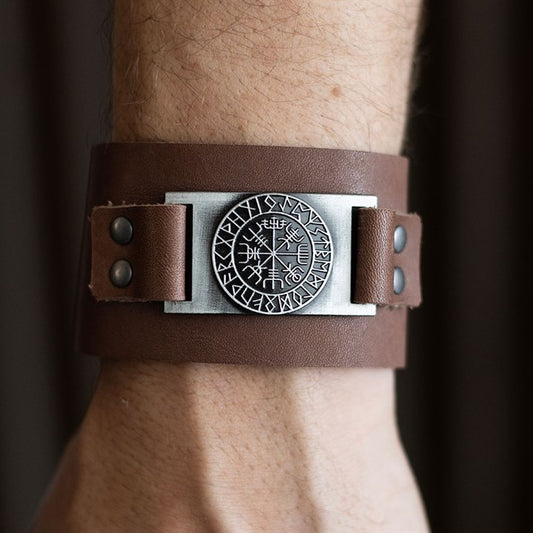 A close-up of a person’s wrist adorned with a wide brown leather cuff Viking bracelet, featuring metal rivets and a circular metal plate engraved with runic symbols and intricate designs.