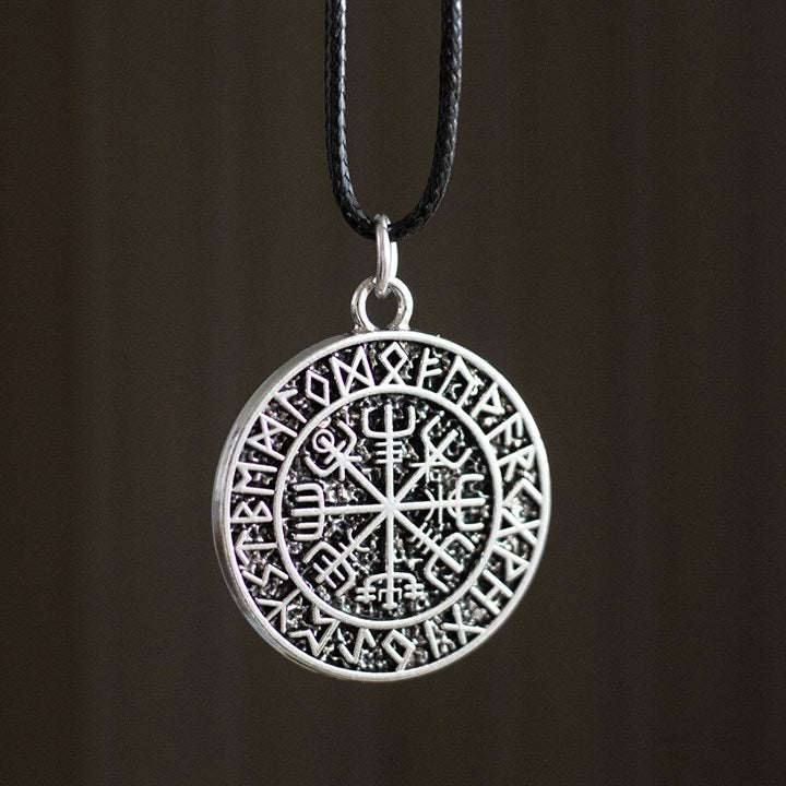 A circular silver Viking necklace pendant adorned with Norse runes around the rim and a central Vegvisir Viking Compass, renowned for its protective symbolism, hangs from a slender black cord against a blurred brown backdrop.