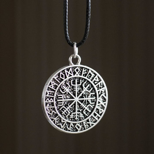 A circular silver Viking necklace pendant adorned with Norse runes around the rim and a central Vegvisir Viking Compass, renowned for its protective symbolism, hangs from a slender black cord against a blurred brown backdrop.
