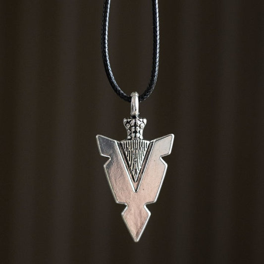 A silver arrowhead necklace with engraved detailing hangs from a black cord against a dark background. The pendant displays geometric lines, a textured upper section, and echoes of protective symbolism inspired by Viking jewellery.