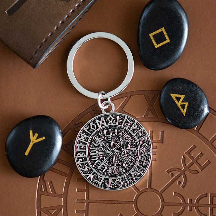 A metal Viking compass keyring with a round pendant engraved with Norse runes lies on a brown surface, surrounded by three black stones featuring yellow runic symbols and a partial brown leather wallet in the top left corner.
