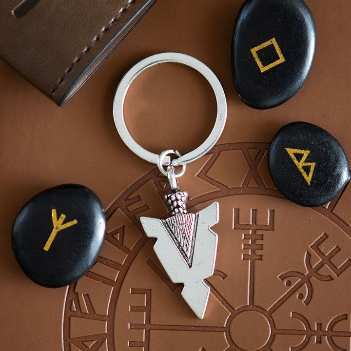 A silver arrowhead keyring with a geometric pendant lies on a brown surface adorned with Norse designs, evoking Viking heritage. Three black stones painted with yellow runes and a brown wallet complete the scene.
