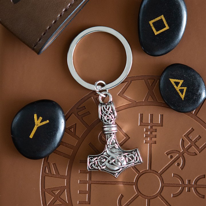 A silver Mjölnir hammer keyring rests on a brown surface bearing a Norse symbol, surrounded by three black stones with yellow runes, evoking Viking heritage. A leather wallet is partly visible in the top left corner.