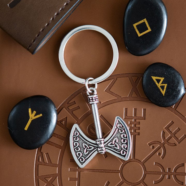 A silver Axe of Valour keyring, shaped like a double-headed axe, rests on a brown surface with engraved Norse symbols. It is surrounded by three black stones bearing yellow runes and a brown leather item in the corner.