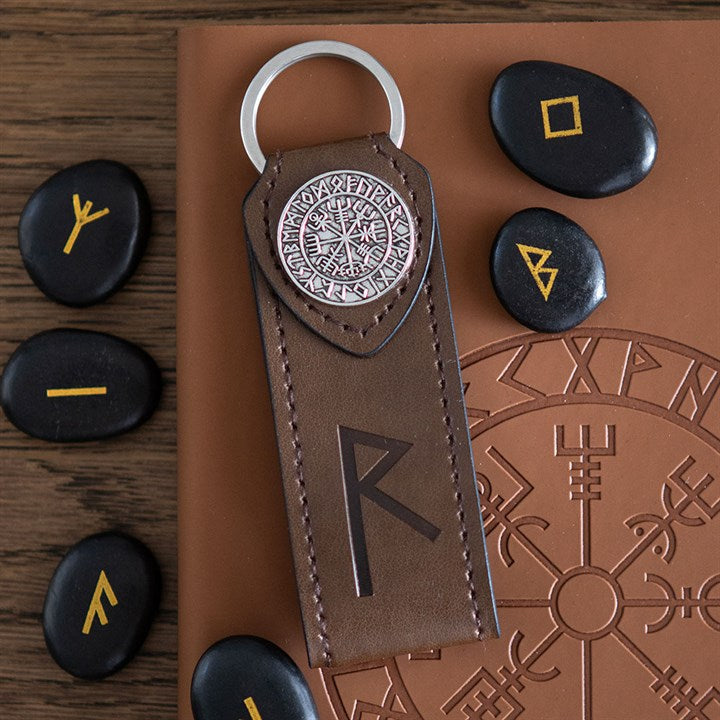 A brown Viking rune keyring featuring a Nordic rune and metal emblem lies on a notebook engraved with runes. Black stones inscribed with yellow runes are scattered on a wooden surface—a unique gift for travellers.