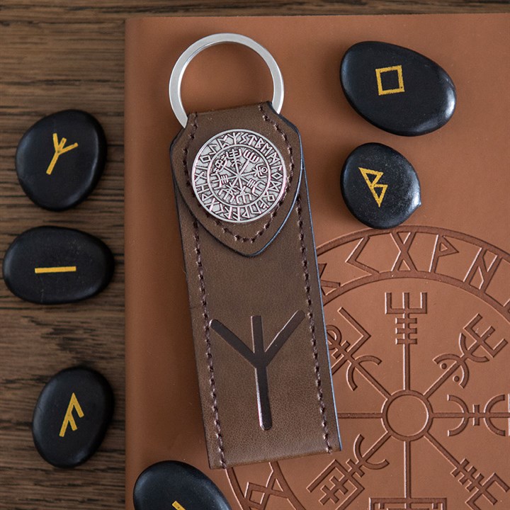 A brown leather protection keyring with Norse runes and a Vegvisir symbol lies on a journal adorned with runic designs. Several black stones marked with yellow runes encircle the keyring and journal on a wooden surface.