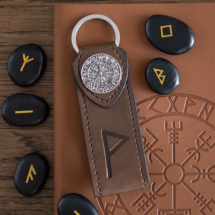 A brown leather Viking rune keyring with a metal emblem lies atop a notebook featuring a Viking symbol, surrounded by black rune stones with yellow markings—an ideal gift for travellers interested in Norse heritage.