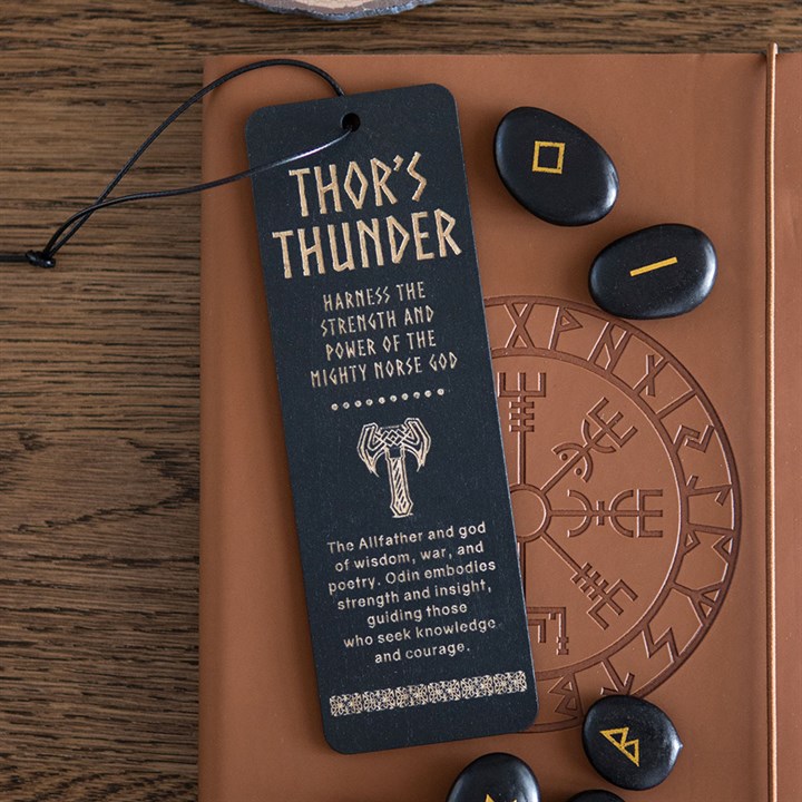 A black wooden bookmark bearing “Thor’s Thunder” in gold lettering rests on a brown book embossed with Nordic symbols. Four black rune stones adorned with gold Norse designs sit nearby on the wooden surface.