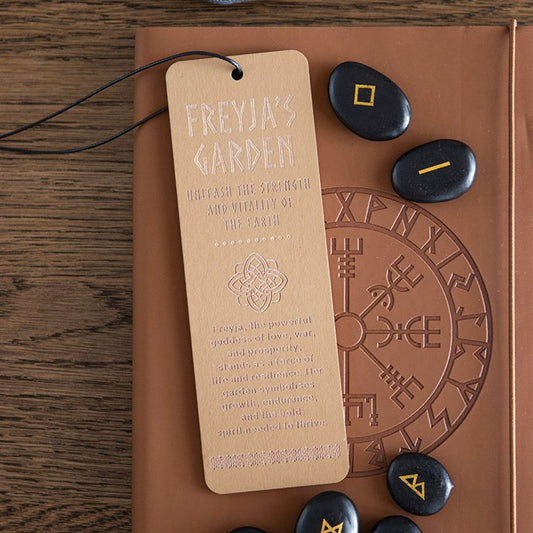 A tan wooden bookmark inscribed with Freyja’s Garden and engraved Norse mythology designs lies on a brown embossed book, surrounded by black rune stones with gold symbols on a wooden surface—a perfect Viking gift.