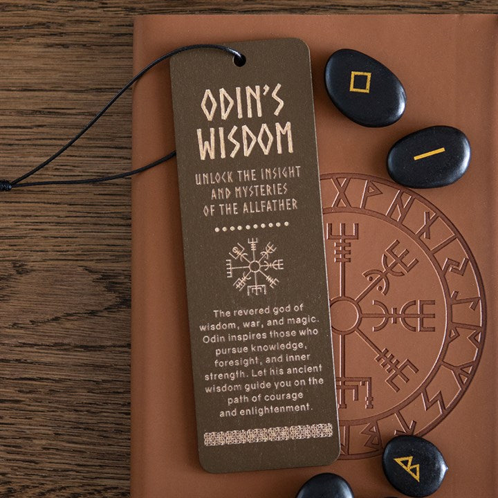 A wooden Viking bookmark bearing Norse symbols and the phrase “Odin’s Wisdom—Unlock the Insight and Mysteries of the Allfather” rests on a runic journal beside four black runestones with yellow and orange symbols, atop a wooden table.