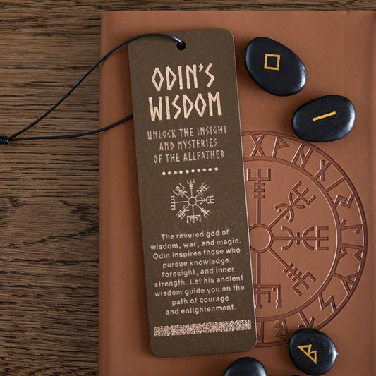 A wooden Viking bookmark bearing Norse symbols and the phrase “Odin’s Wisdom—Unlock the Insight and Mysteries of the Allfather” rests on a runic journal beside four black runestones with yellow and orange symbols, atop a wooden table.