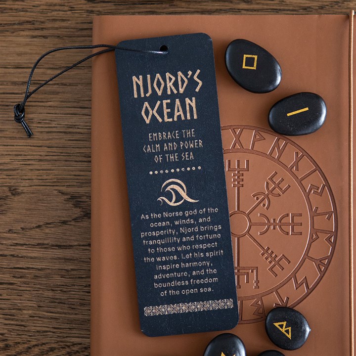 A black Njord’s Ocean wooden bookmark inscribed “Embrace the calm and power of the sea” rests on a brown embossed book, with rune stones close by—ideal for sea lovers and admirers of the Norse god of the ocean.