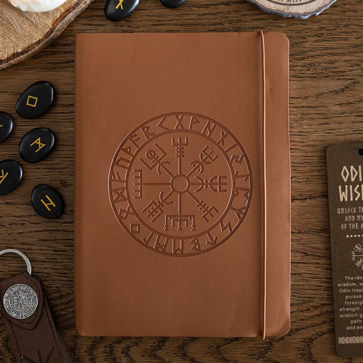 A brown faux leather notebook embossed with a circular Norse symbol and runic alphabet rests on a wooden table. Nearby are rune stones, a keyring, a card, and a wooden tray filled with additional runes.