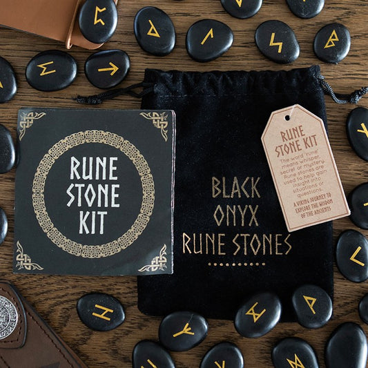A Rune Stone Kit featuring a decorative box, tag, black velvet pouch labelled “BLACK ONYX RUNE STONES,” and several black onyx Viking rune stones engraved with gold Norse runes scattered across a wooden surface.