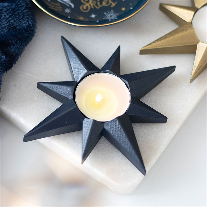 A black, star-shaped candle holder with a lit tealight sits on white marble, next to a gold star candle holder and a blue dish. The celestial décor glows warmly, creating a cosy ambience.