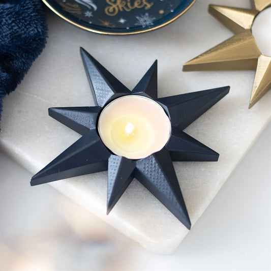 A black, star-shaped candle holder with a lit tealight sits on white marble, next to a gold star candle holder and a blue dish. The celestial décor glows warmly, creating a cosy ambience.