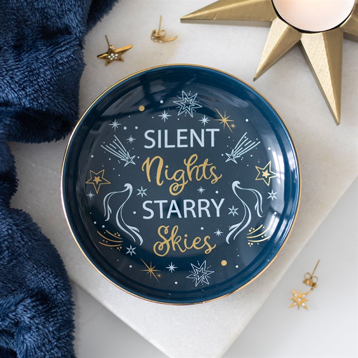A celestial-inspired ceramic dish with gold-edged detailing features “Silent Nights Starry Skies” in white and gold script, surrounded by cosmic charm—stars, comets, and shooting stars. Blue fabric and gold star decorations enhance the night sky atmosphere.