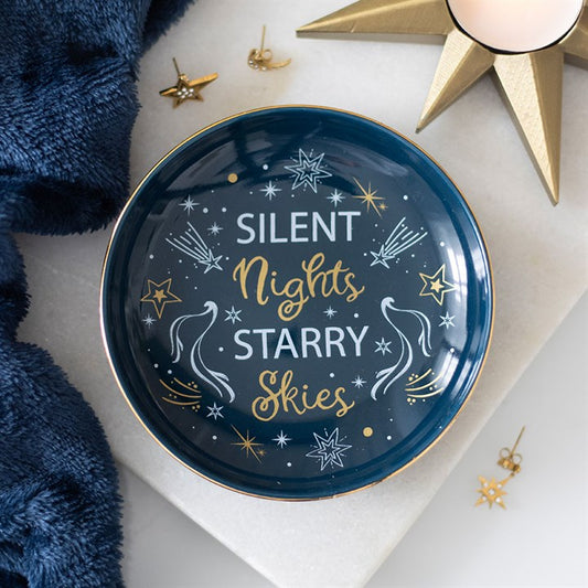 A celestial-inspired ceramic dish with gold-edged detailing features “Silent Nights Starry Skies” in white and gold script, surrounded by cosmic charm—stars, comets, and shooting stars. Blue fabric and gold star decorations enhance the night sky atmosphere.