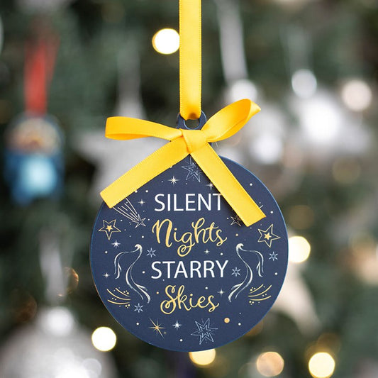 A round navy Christmas tree bauble adorned with yellow stars, white swirls, and the words “Silent Nights Starry Skies” in white and yellow hangs from a yellow ribbon, adding celestial sparkle to a blurred Christmas tree backdrop.
