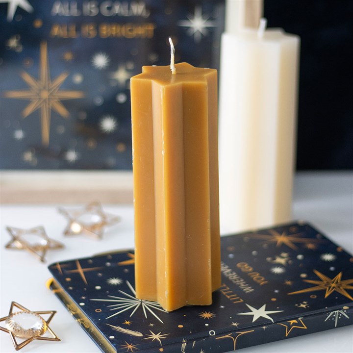 A tall, hexagonal, honey-coloured festive candle stands upright on a dark book adorned with gold star patterns. A cream-coloured candle and additional star-themed décor appear softly blurred in the background. The scene is both festive and elegant.