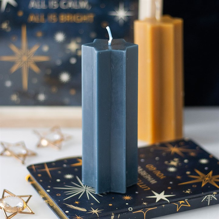 A tall, hexagonal navy candle stands on a star-patterned book, with a gold candle and decorative star lights blurred in the background—a perfect festive table decoration with celestial motifs and a sign reading ALL IS CALM, ALL IS BRIGHT.