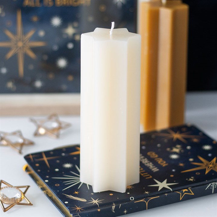 A tall, white, unscented candle stands on a dark blue book patterned with stars. In the background are a honey-coloured candle, star-shaped ornaments, and a dark box adorned with gold stars and the phrase “ALL IS BRIGHT.”.