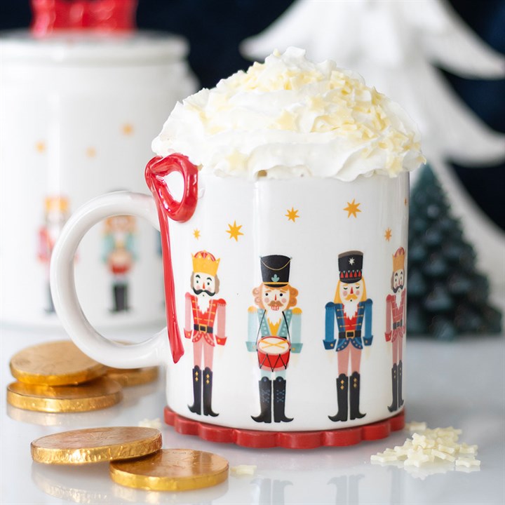 A festive mug from the Christmas mug collection, decorated with nutcracker soldiers, holds a hot drink topped with whipped cream and white chocolate shavings. Gold coins and a white bauble add festive charm in the background.