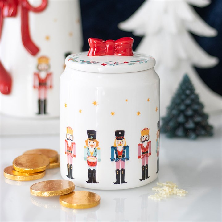 A ceramic storage jar featuring a charming nutcracker design and a red bow on the lid sits on a table, surrounded by gold-wrapped chocolate coins, star-shaped sprinkles, and festive Christmas decorations in the background.