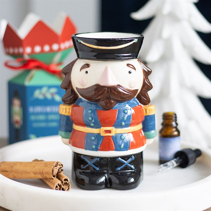 A colourful ceramic mug shaped like a Nutcracker soldier stands on a marble tray, with cinnamon sticks, a small brown bottle with a dropper, and the perfect Christmas present and white Christmas tree in the background.