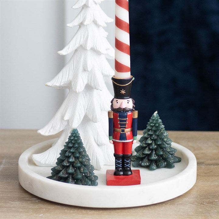 A decorative scene featuring a nutcracker-shaped figurine, two green pine tree miniatures, a white Christmas tree, and a red-and-white striped candle on a round white tray atop a wooden surface adds Christmas charm.
