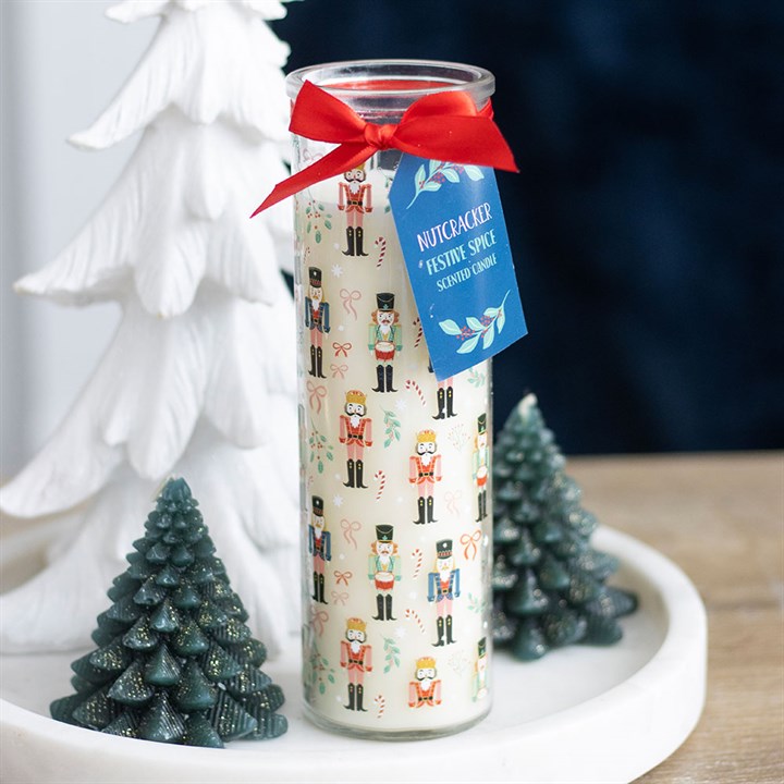 A tall Christmas candle in a glass holder with nutcracker design and candy canes rests on a white tray, surrounded by festive spice scents, green baubles, and a white decorative tree.
