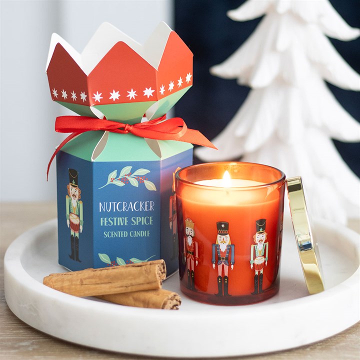 A nutcracker candle sits on a white tray with cinnamon sticks. Next to it, a colourful nutcracker-themed box with a red bow adds to the seasonal décor. A white decorative tree is blurred in the background.