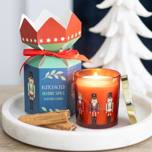 A nutcracker candle sits on a white tray with cinnamon sticks. Next to it, a colourful nutcracker-themed box with a red bow adds to the seasonal décor. A white decorative tree is blurred in the background.