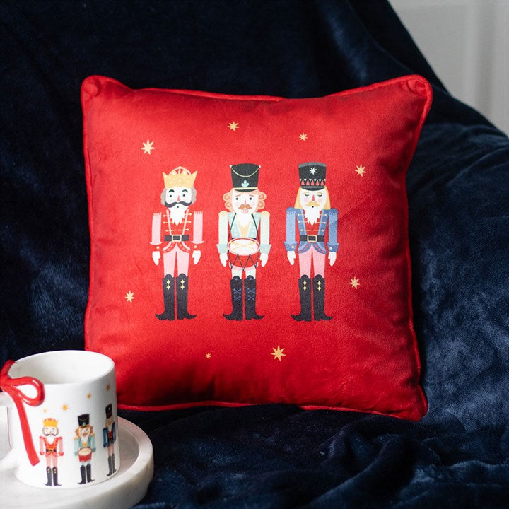A red Christmas cushion decorated with three illustrated nutcrackers rests against a dark blue blanket. Nearby, a white mug featuring nutcracker designs and a red spoon adds extra festive charm to the scene.