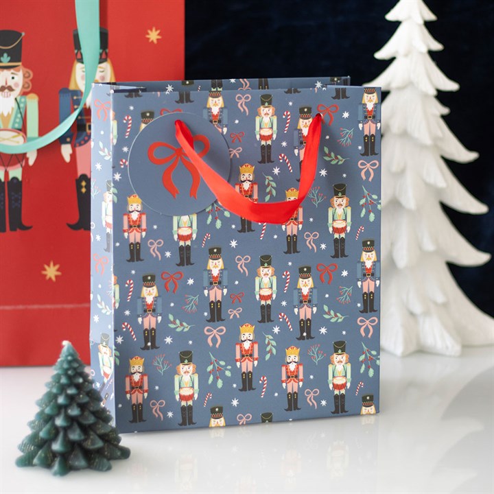 A festive, nutcracker-themed blue Christmas gift bag adorned with candy cane and holly patterns, red ribbon handles, and a round tag sits between green and white decorative trees—perfect for Secret Santa surprises.
