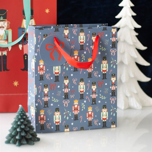 A festive, nutcracker-themed blue Christmas gift bag adorned with candy cane and holly patterns, red ribbon handles, and a round tag sits between green and white decorative trees—perfect for Secret Santa surprises.