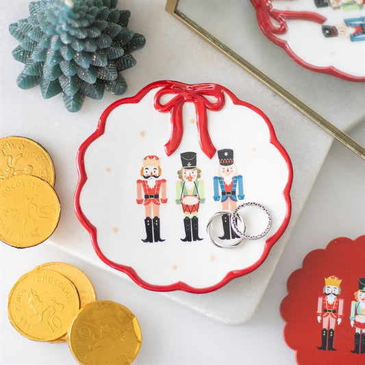 A ceramic trinket dish with scalloped edges, a red rim and a bow holds two rings and features three nutcracker figures. Nearby are gold chocolate coins, a green decorative tree and part of a mirror reflecting the dish.