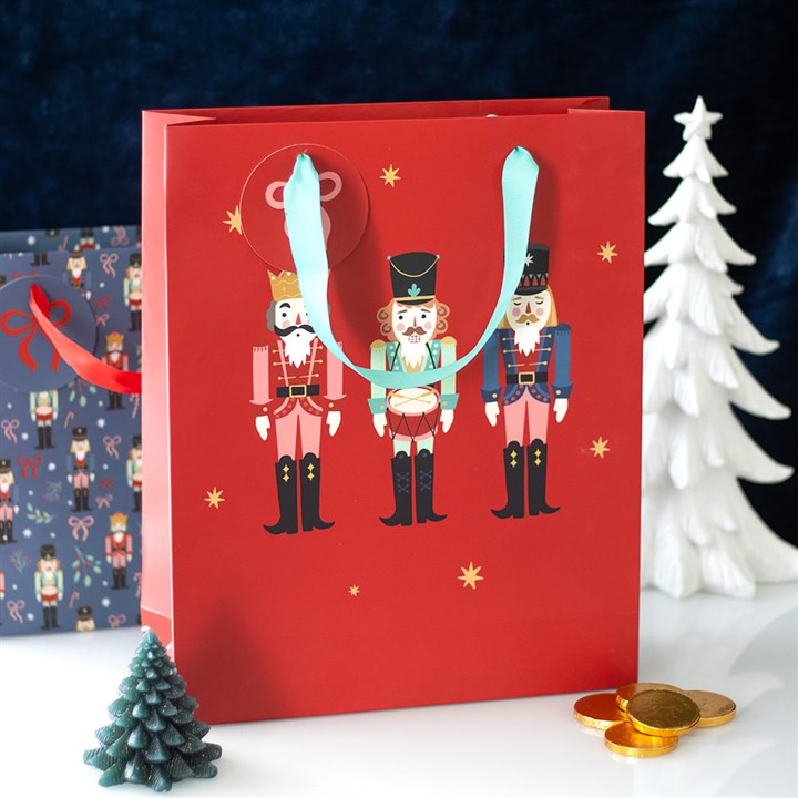 A festive red Christmas gift bag with a nutcracker design and light blue handles sits beside a white tree, a small green tree, gold coins, and a dark gift bag with a nutcracker pattern on a white surface.