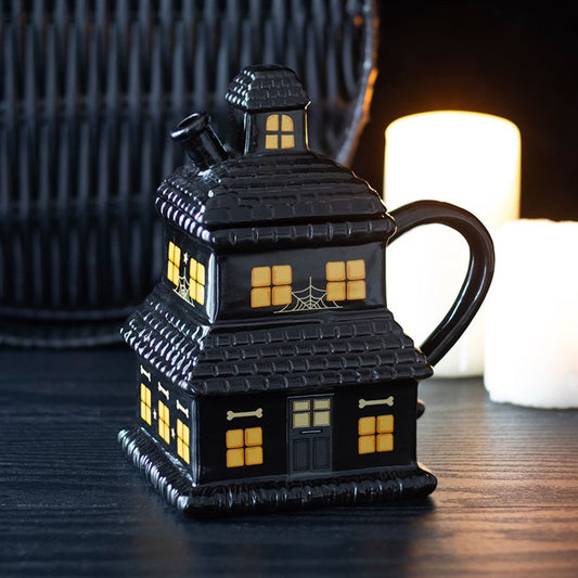 A ceramic teapot shaped like a haunted house, featuring glowing yellow windows, a crooked chimney, and spiderweb details, sits on a dark table, creating an eerie scene perfect for any Halloween mug or ceramic mug collection.