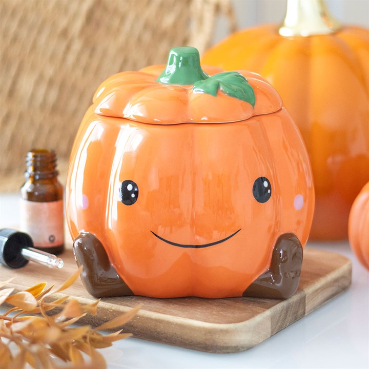 A ceramic, pumpkin-shaped wax warmer with a smiling face and rosy cheeks sits on a wooden board. The container features a green stem lid and brown feet, making it perfect for adding Halloween charm to your décor.