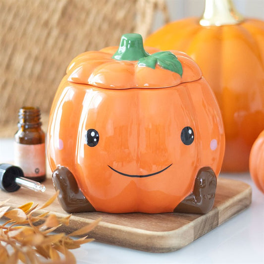 A ceramic, pumpkin-shaped wax warmer with a smiling face and rosy cheeks sits on a wooden board. The container features a green stem lid and brown feet, making it perfect for adding Halloween charm to your décor.