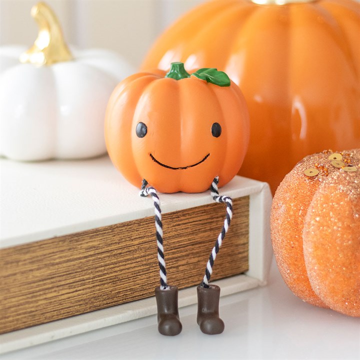 A small, smiling pumpkin ornament with black eyes, rope legs, and brown boots sits on a book. Larger decorative pumpkins, perfect for Hallowe’en décor, are displayed in the background.