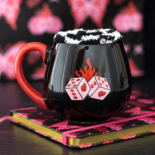 A quirky present: this black mug with a red handle—flaming dice with heart motifs—rests on a notebook decorated with pink butterflies. Black and white whipped cream imitates chequerboard socks, mixing fun and charm.