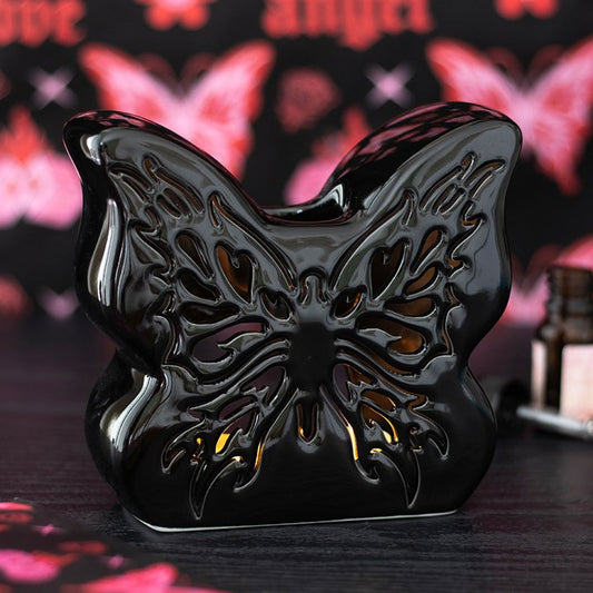 A glossy black ceramic butterfly oil burner with intricate cut-outs rests on a dark surface, ready for a tealight and fragrance oil. Pink and red butterfly and love designs appear blurred in the background.