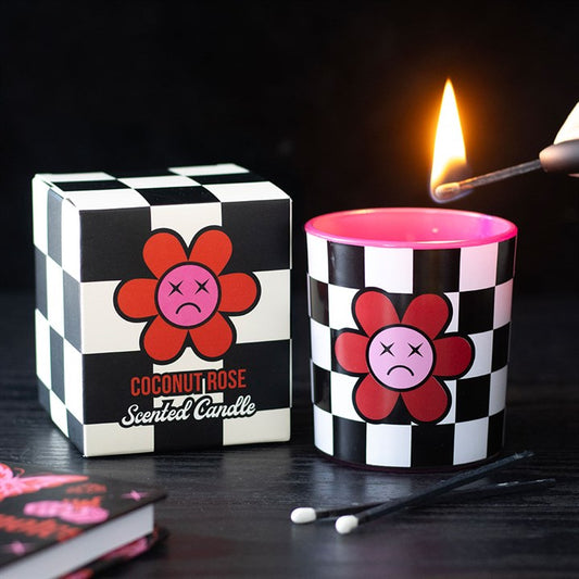 A hand lights a chequerboard candle with a pink interior and a sad flower design; its box reads “Coconut Rose Scented Candle.” Cotton buds rest nearby.