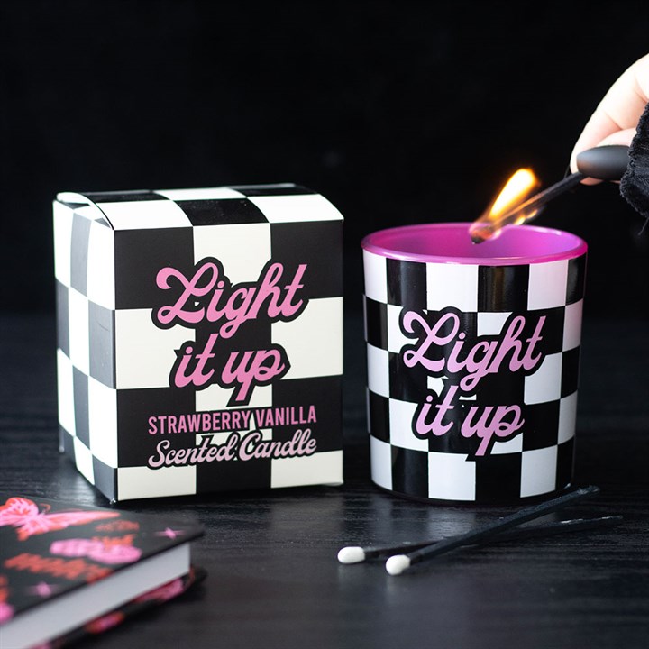 A hand lights a match above a pop-punk chequerboard candle featuring “Light it up” in pink script. The matching strawberry vanilla box sits nearby, with two unused matches on the black surface.