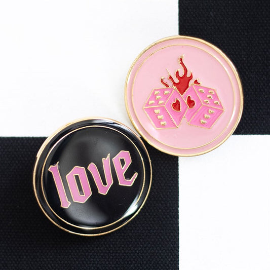 Two round enamel badges on a black and white chequered background. This badge set features “love” in pink Gothic lettering and bold pink flaming dice with red hearts—perfect for Y2K rebellion.
