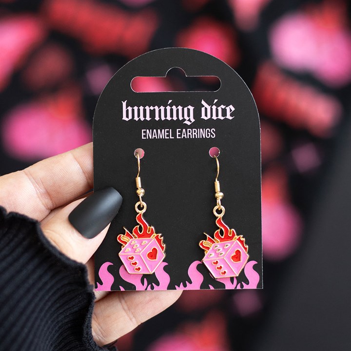 A hand with black matte nails holds a card labelled “burning dice ENAMEL EARRINGS,” showing pink and red earrings—dice with flames and hearts—ideal for alternative or rebellious looks, against a blurred pink-black background.